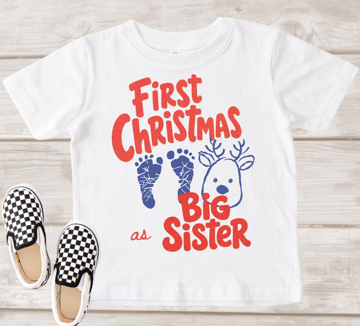 A white short-sleeve kids' T-shirt laid flat on a light wood background, printed with the phrase 'First Christmas as Big Sister' in red lettering, blue baby footprints, and a blue reindeer face. Black-and-white check slip-on shoes are placed beside the shirt as styling props.