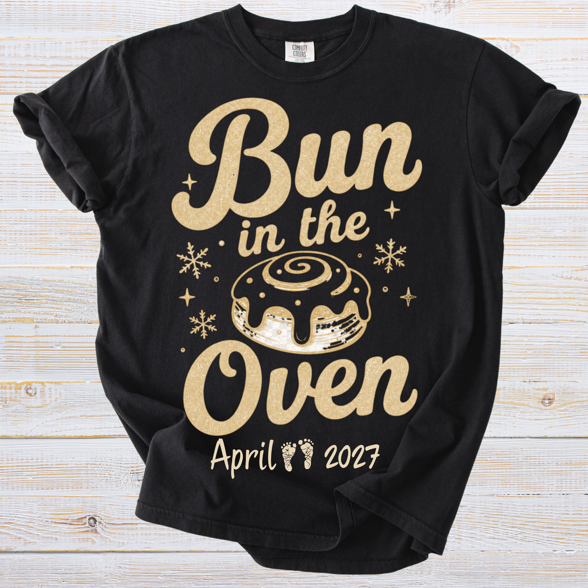 A black short-sleeve t-shirt with a gold glitter-style graphic that reads 'Bun in the Oven' with a cinnamon bun illustration, snowflakes, and date near the hem, shown on a light wood background.