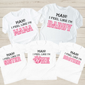 Five white graphic T-shirts arranged on a light wood background, each with different printed family-friendly slogans in black and pink: MAN! I FEEL LIKE I'M MAMA, DADDY, ONE, and BROTHER.