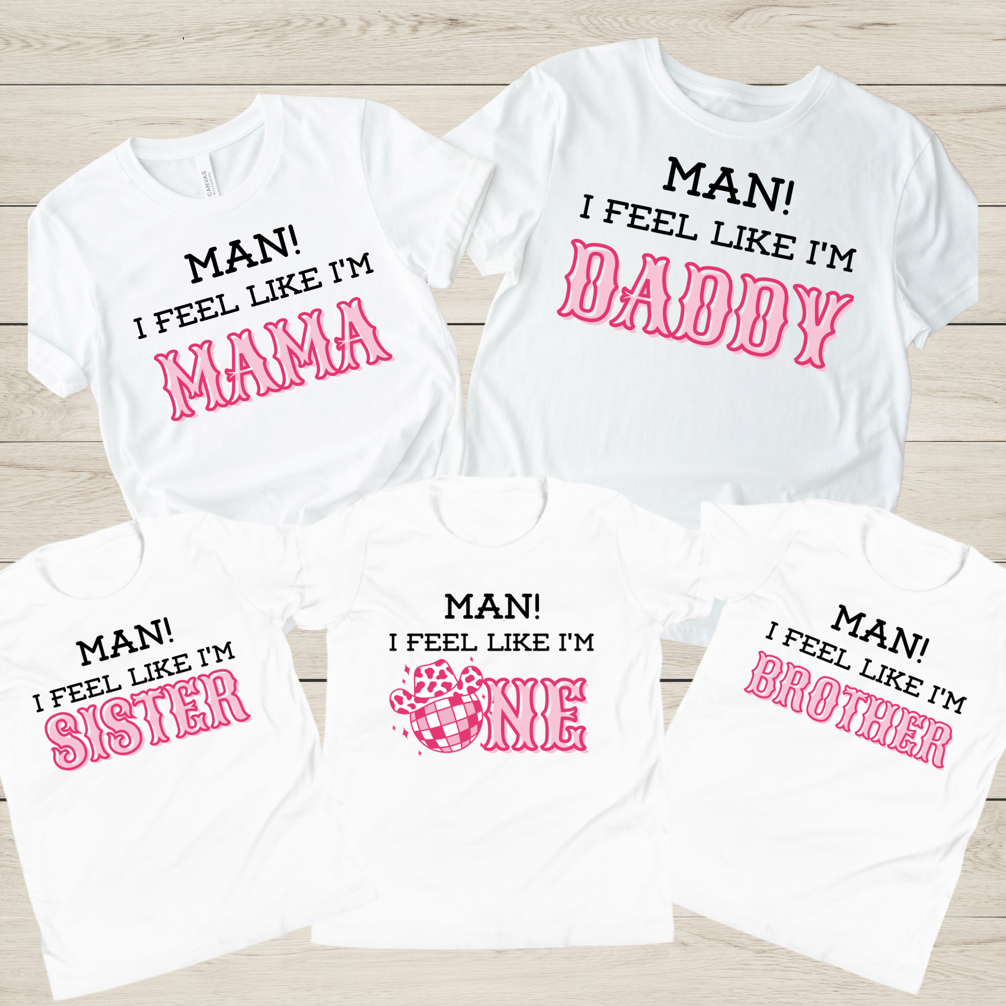Five white graphic T-shirts arranged on a light wood background, each with different printed family-friendly slogans in black and pink: MAN! I FEEL LIKE I'M MAMA, DADDY, ONE, and BROTHER.